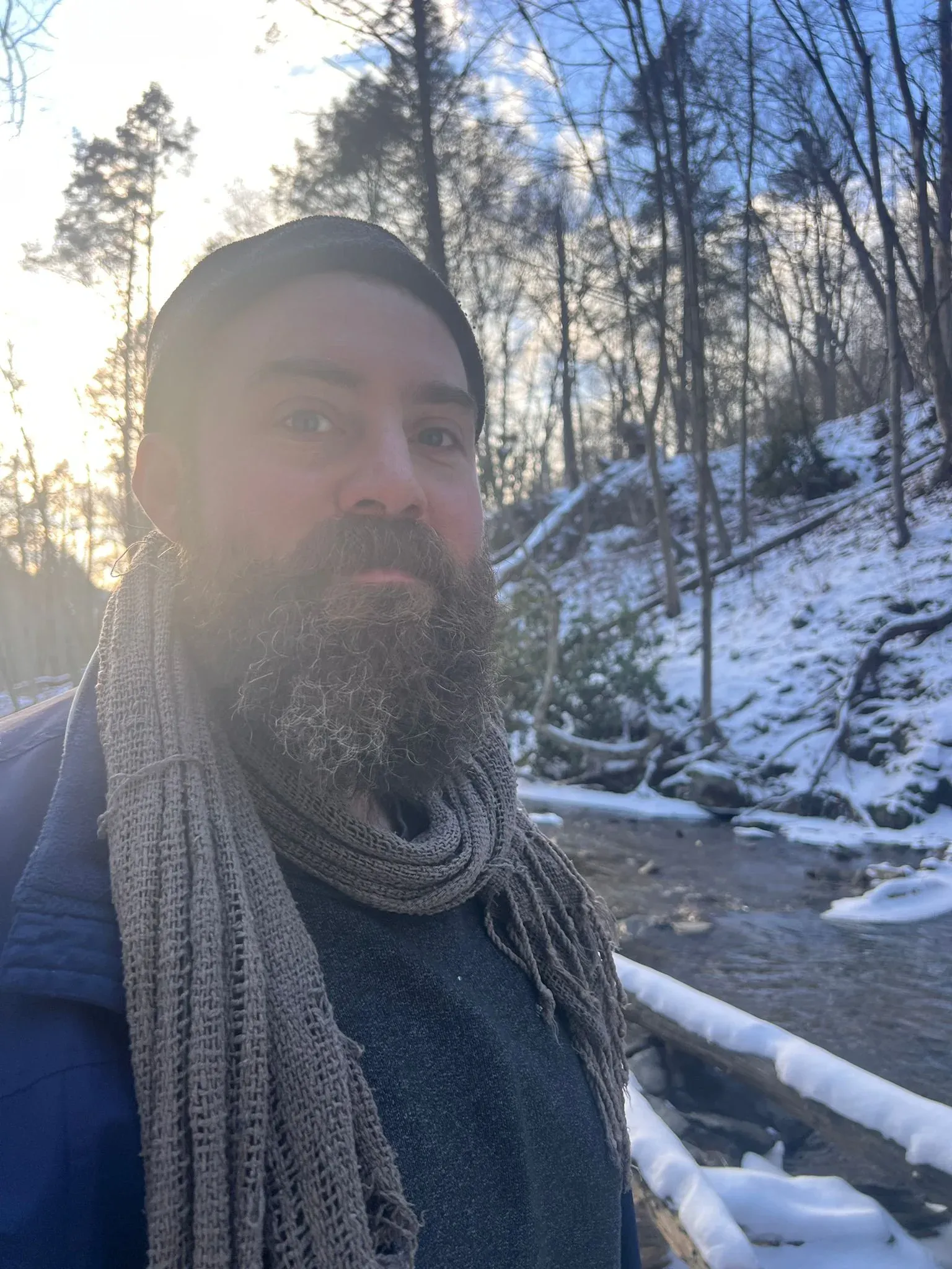 Close portrait of a bearded man in a knitted cap and heavy scarf, snow and low sun behind him.
