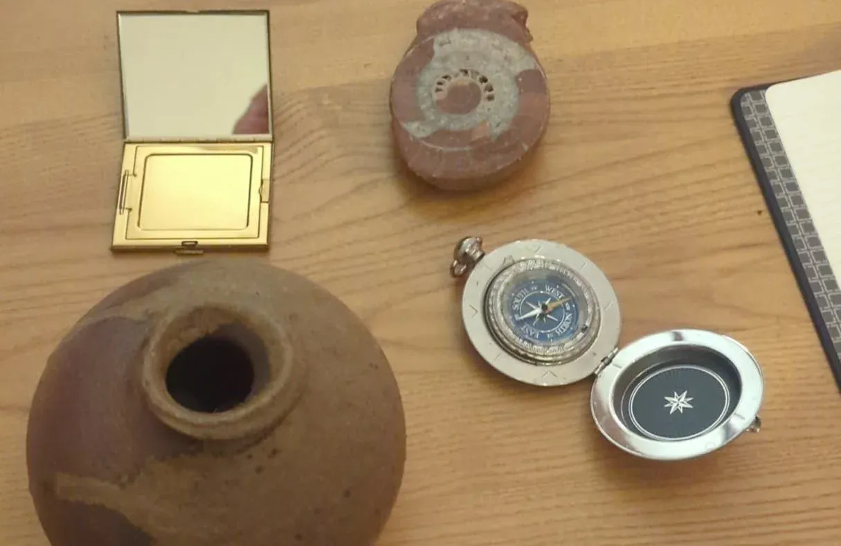 A small earthenware vessel, a folding pocket mirror, and a brass compass laid out on a wooden table beside a closed notebook.