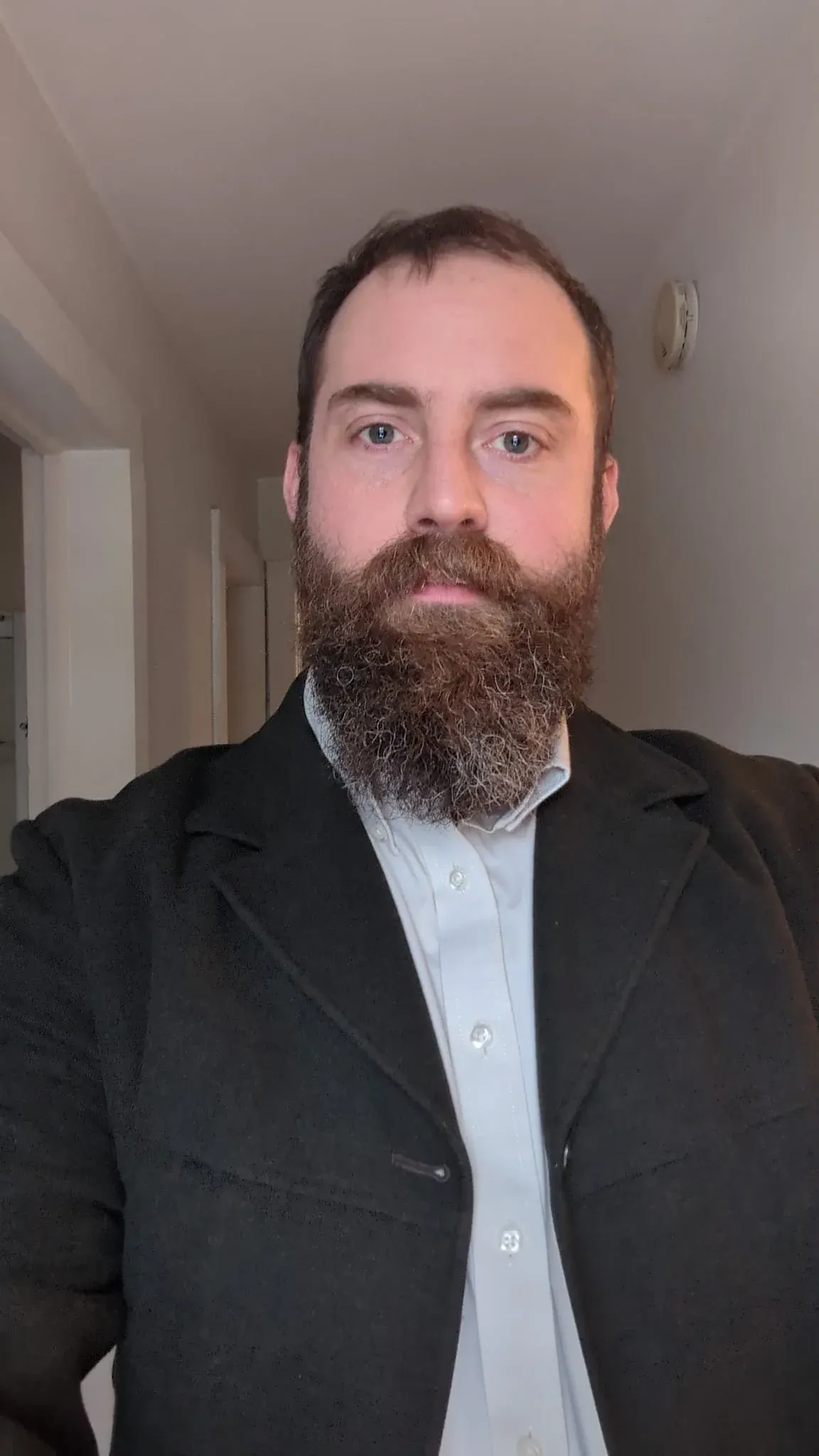 Portrait of a bearded man in a dark jacket and white shirt, standing in a plain hallway.