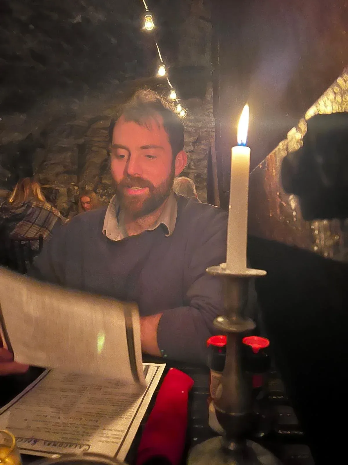 A man reading at a table by the light of a single tall candle, a stone wall behind him.