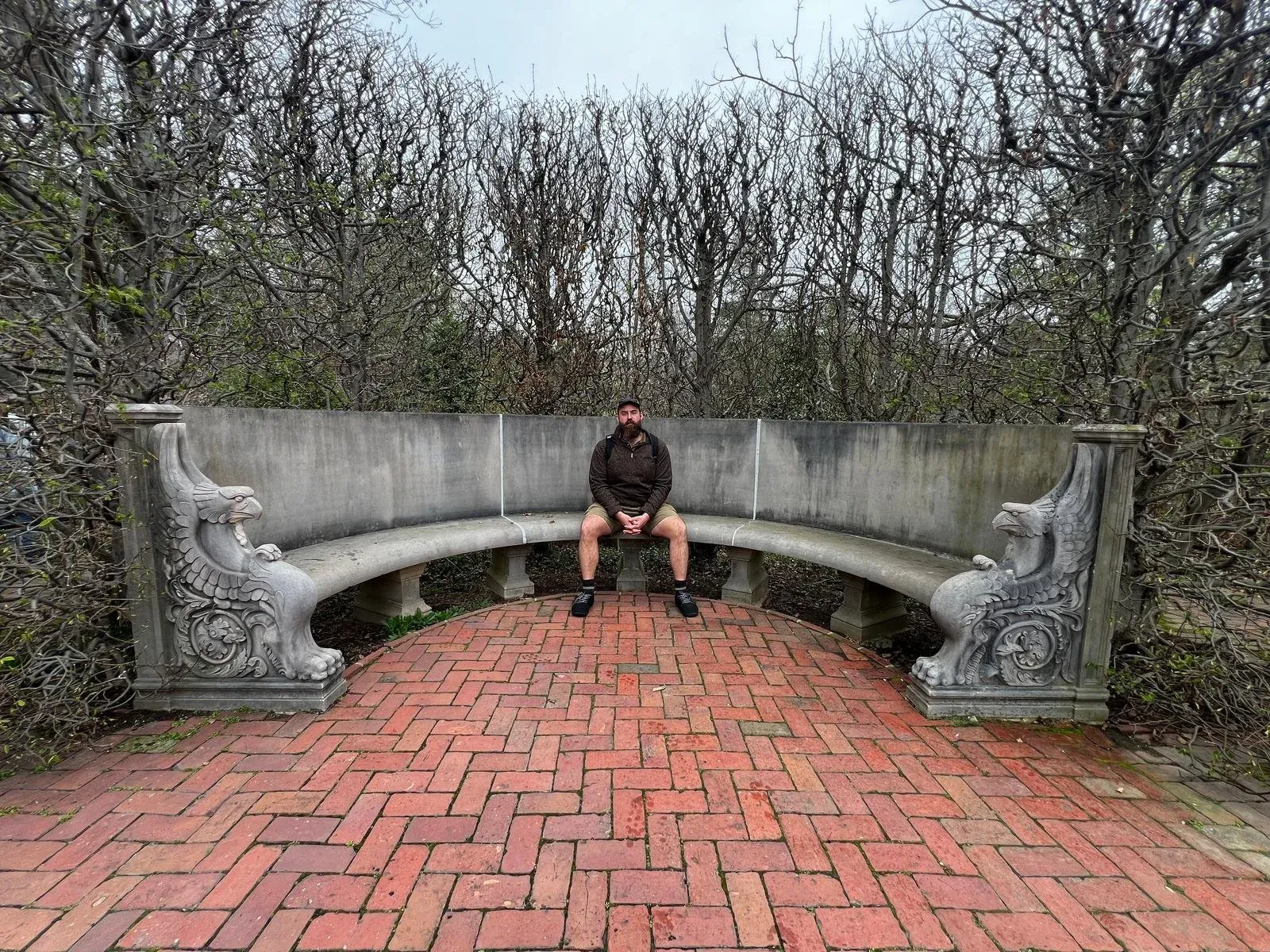 A person sitting on the parapet of a brick footbridge in bare winter woodland.