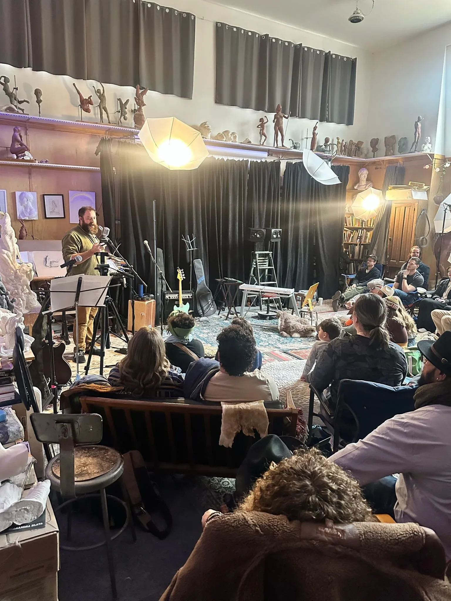 An audience seated in a domestic room turned into a concert space, performers lit at the far end.