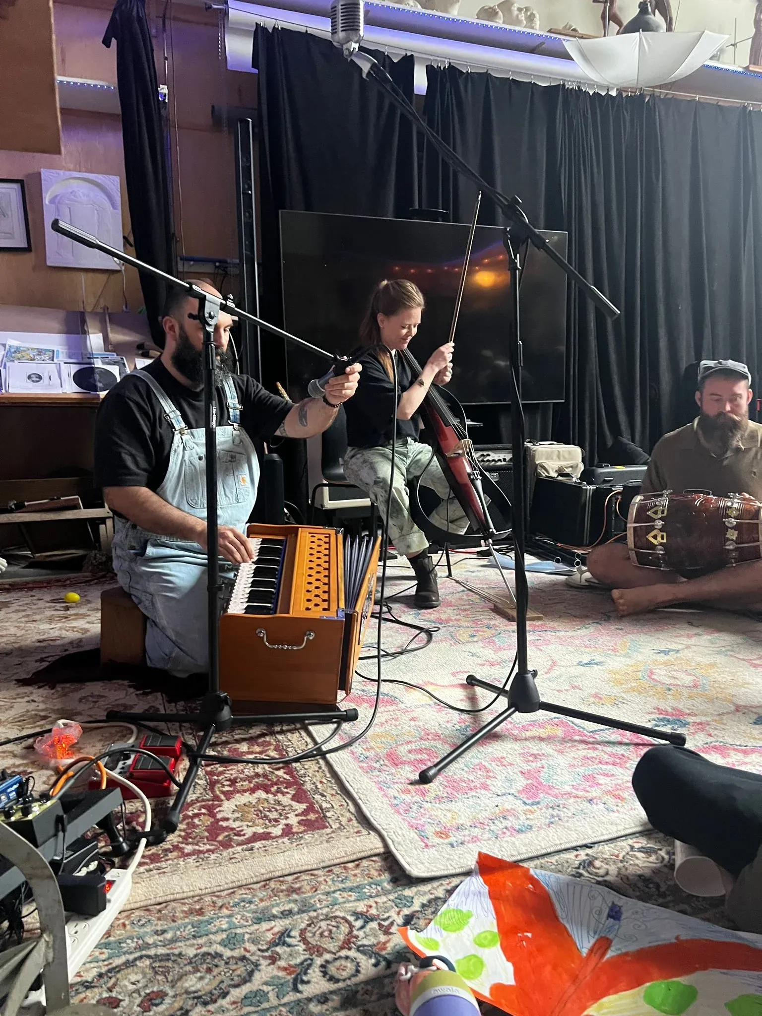Three musicians performing seated on a patterned rug — accordion, voice, and hand drum.