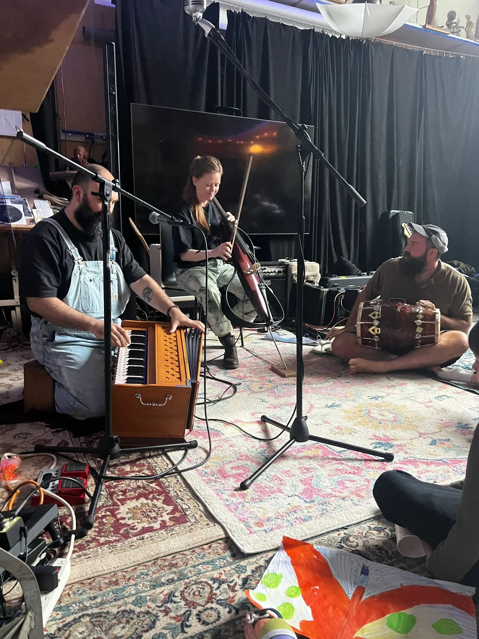 A trio playing on a rug in front of a black curtain, accordion and hand percussion in the foreground.