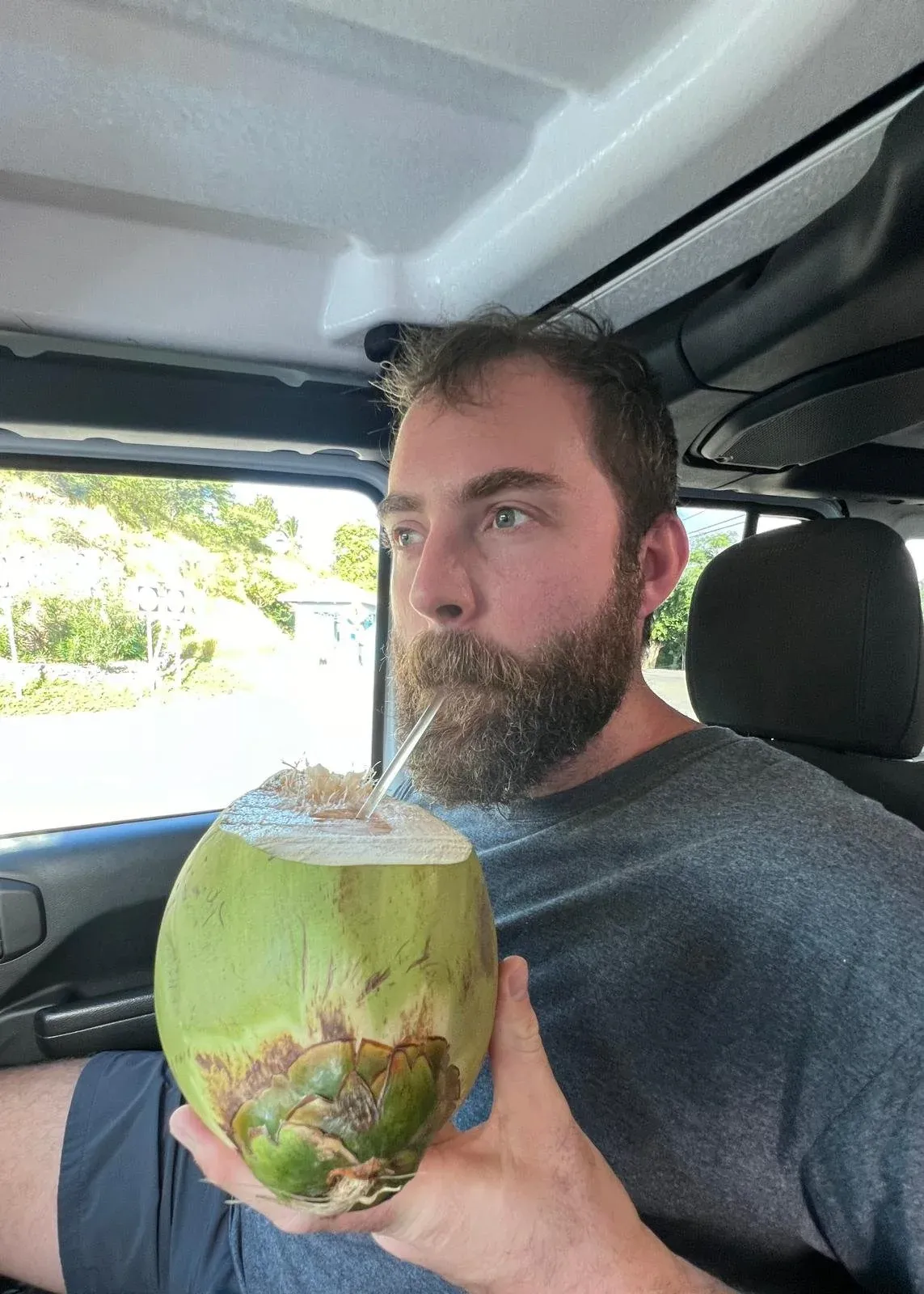 A man drinking from a whole green coconut through a straw, sitting in a parked car.