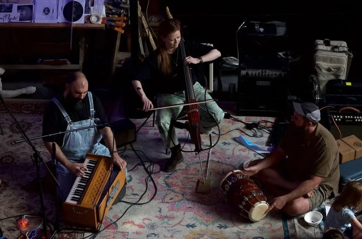 Musicians rehearsing on a rug: keyboard, accordion, and djembe, amplifiers stacked behind.