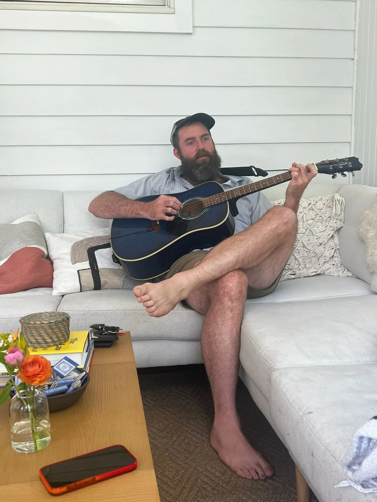 A man playing acoustic guitar barefoot on a white sofa, a low table of flowers in front of him.
