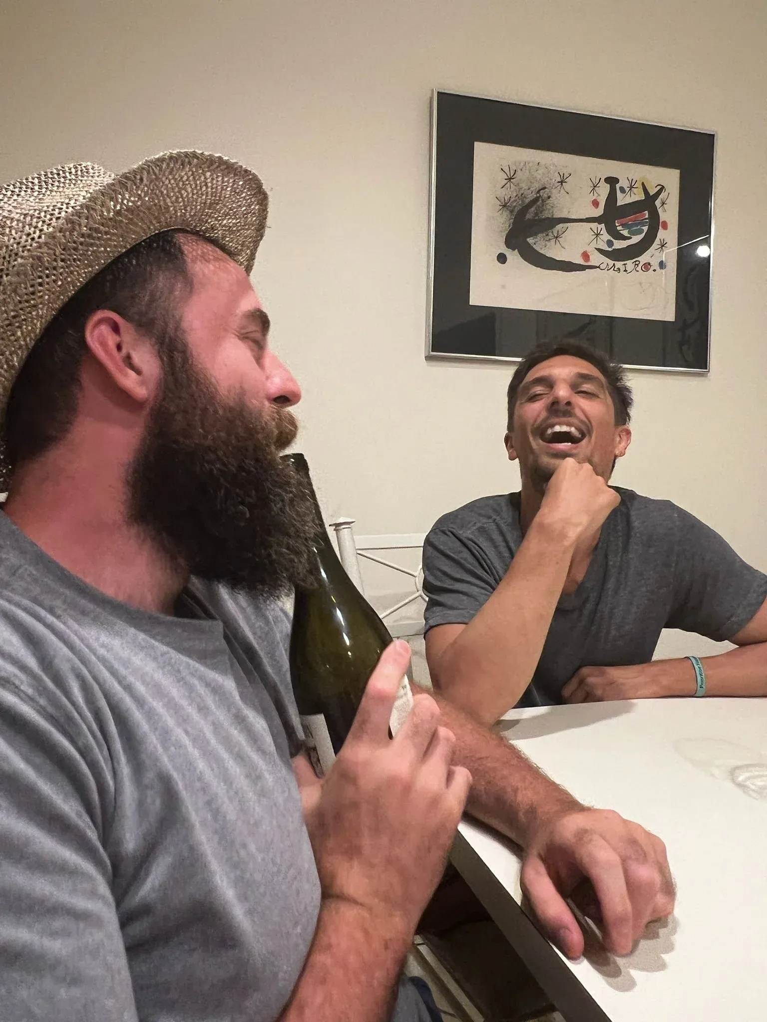 Two men laughing across a kitchen counter, a bottle of wine between them.