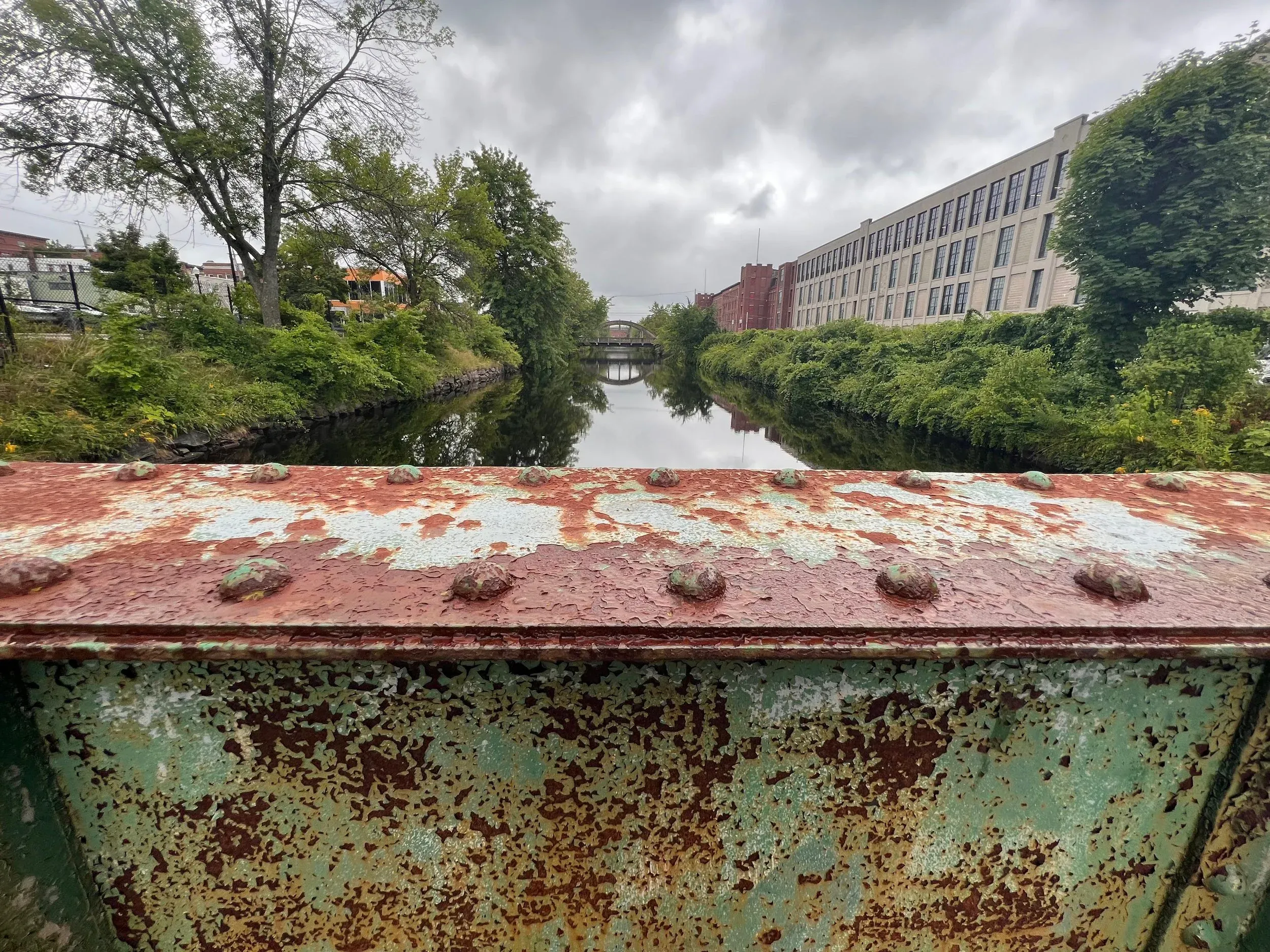 A canal running beside a long brick mill building, seen across a rusted steel edge.