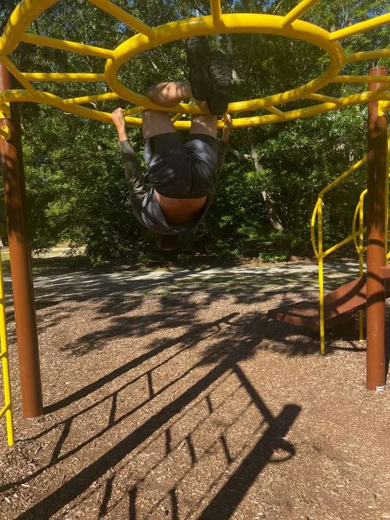 A child hanging upside down from a yellow climbing frame, its shadow thrown across the sand.
