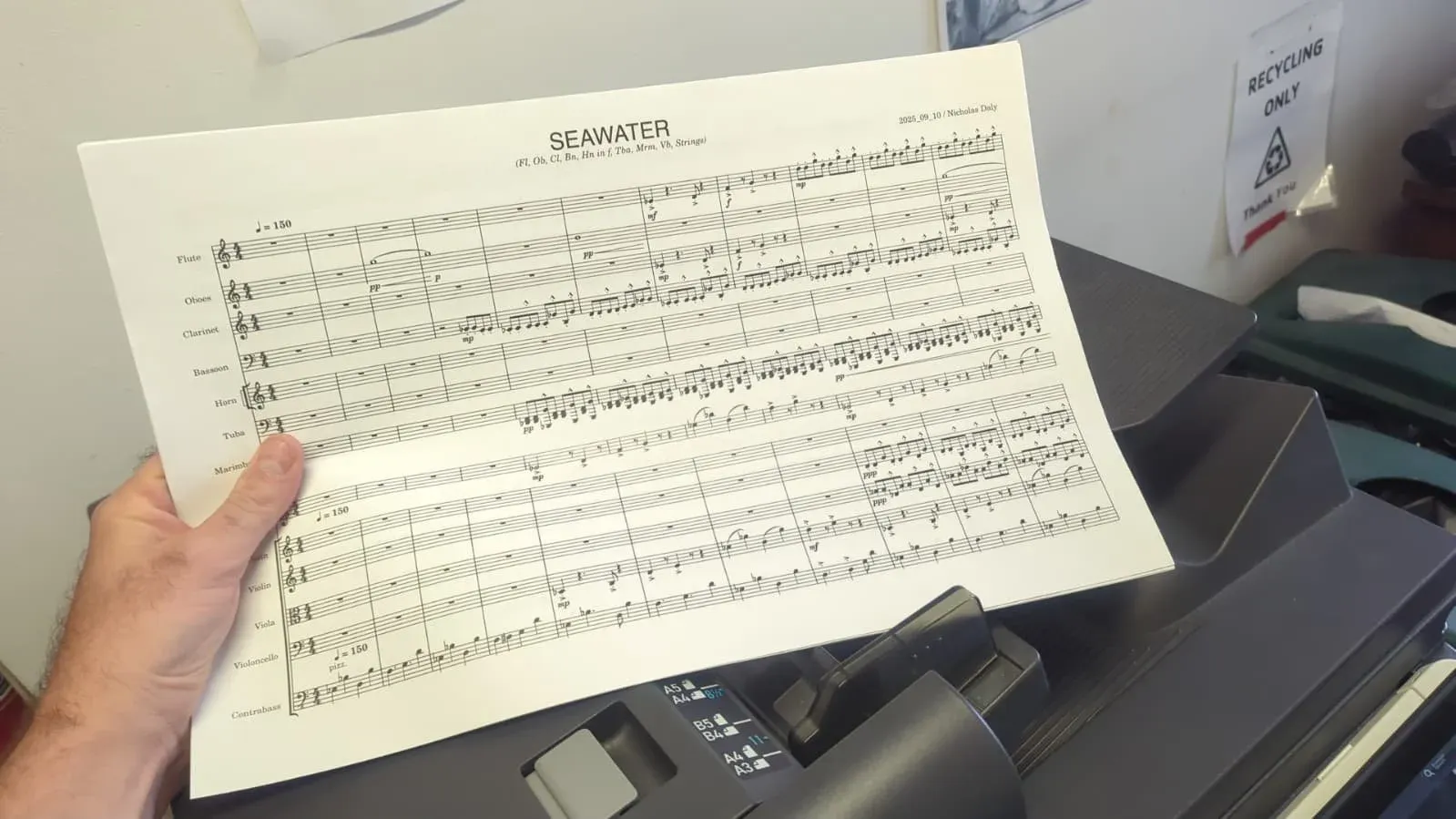 Hands holding a freshly printed page of the SEAWATER score beside a desktop printer.
