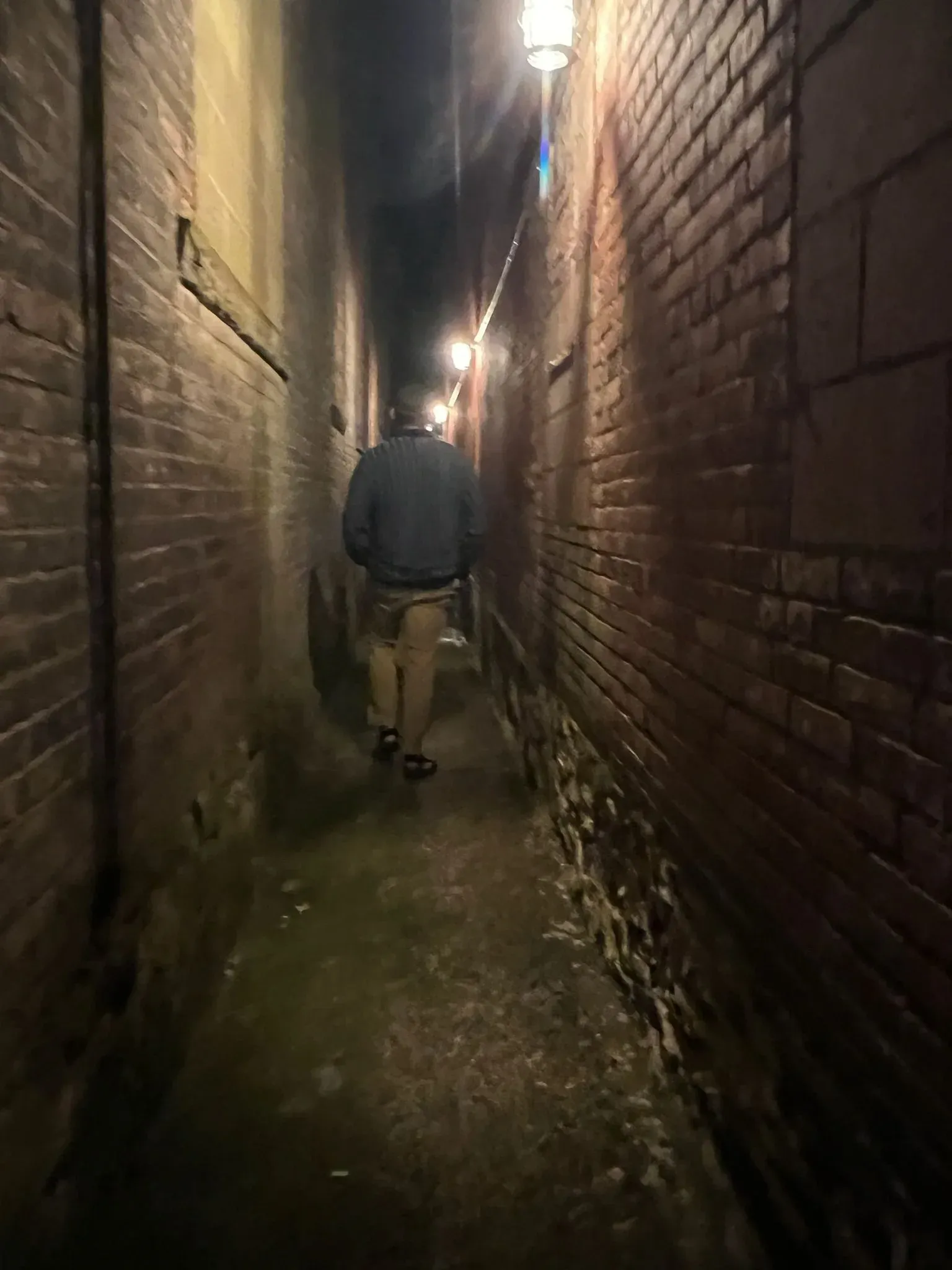 A narrow brick alley lit by a single hanging lamp, a figure walking away into the dark.