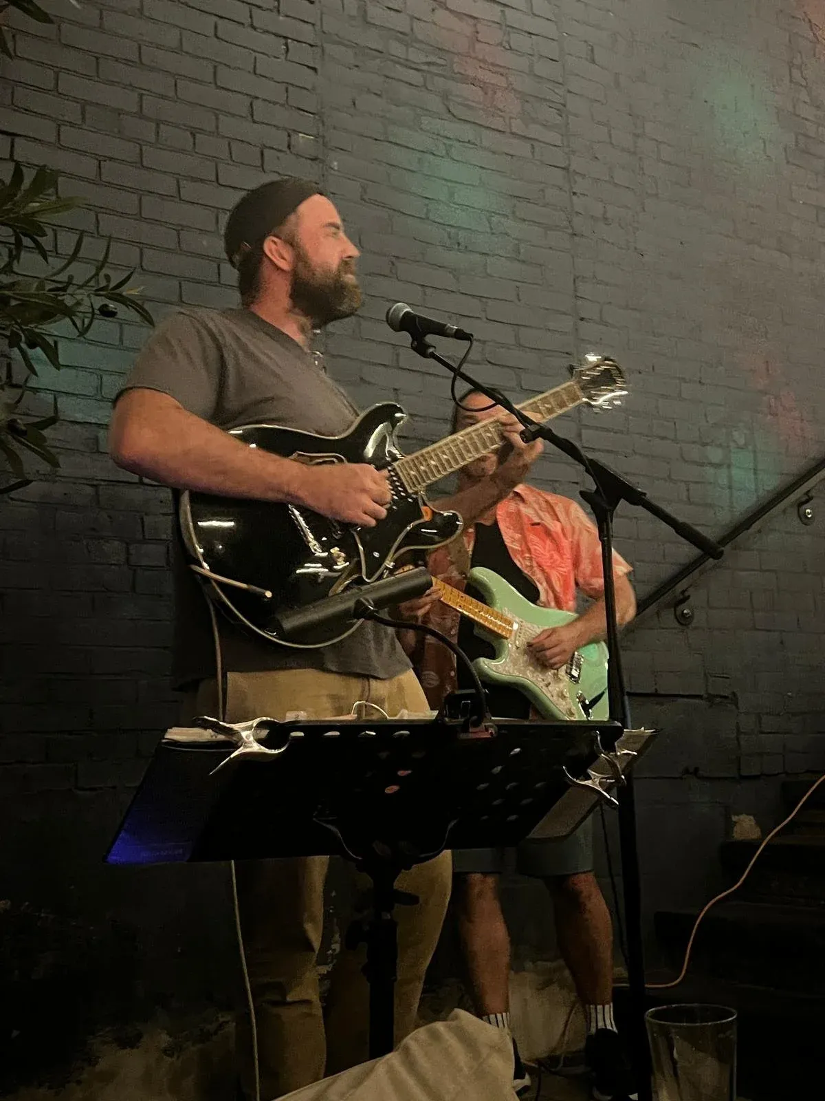 A man playing electric guitar at a microphone in front of a rough stone wall.