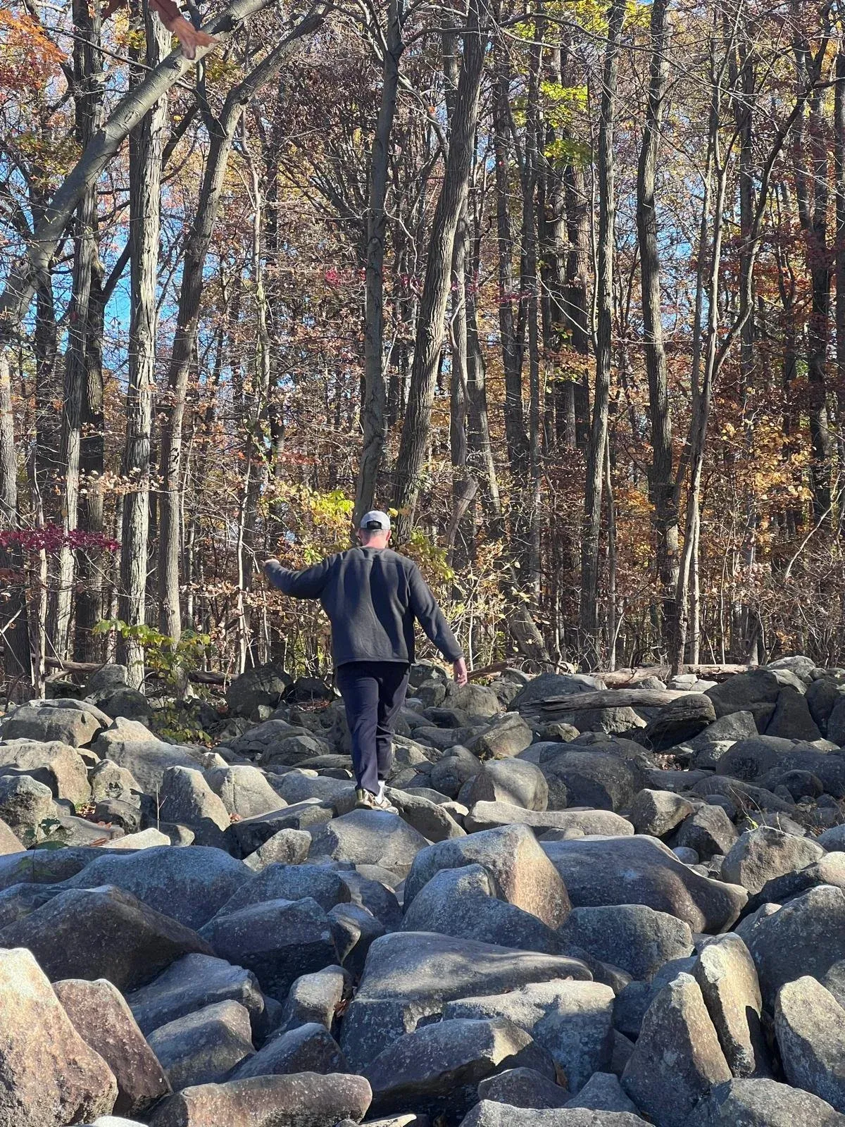 A lone figure standing on a wide boulder field, autumn woods rising behind.