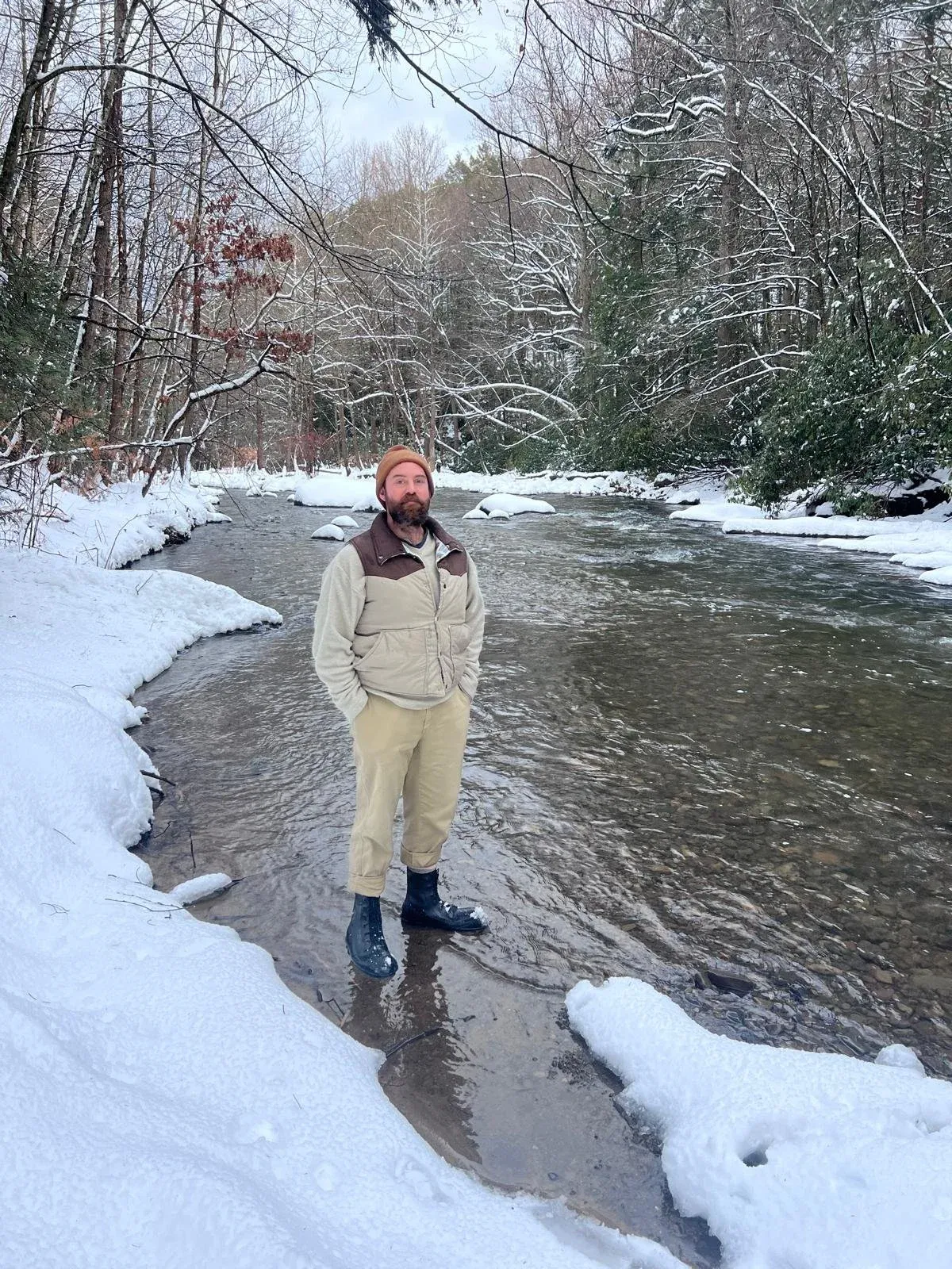 A man standing mid-stream in a shallow creek banked with fresh snow.
