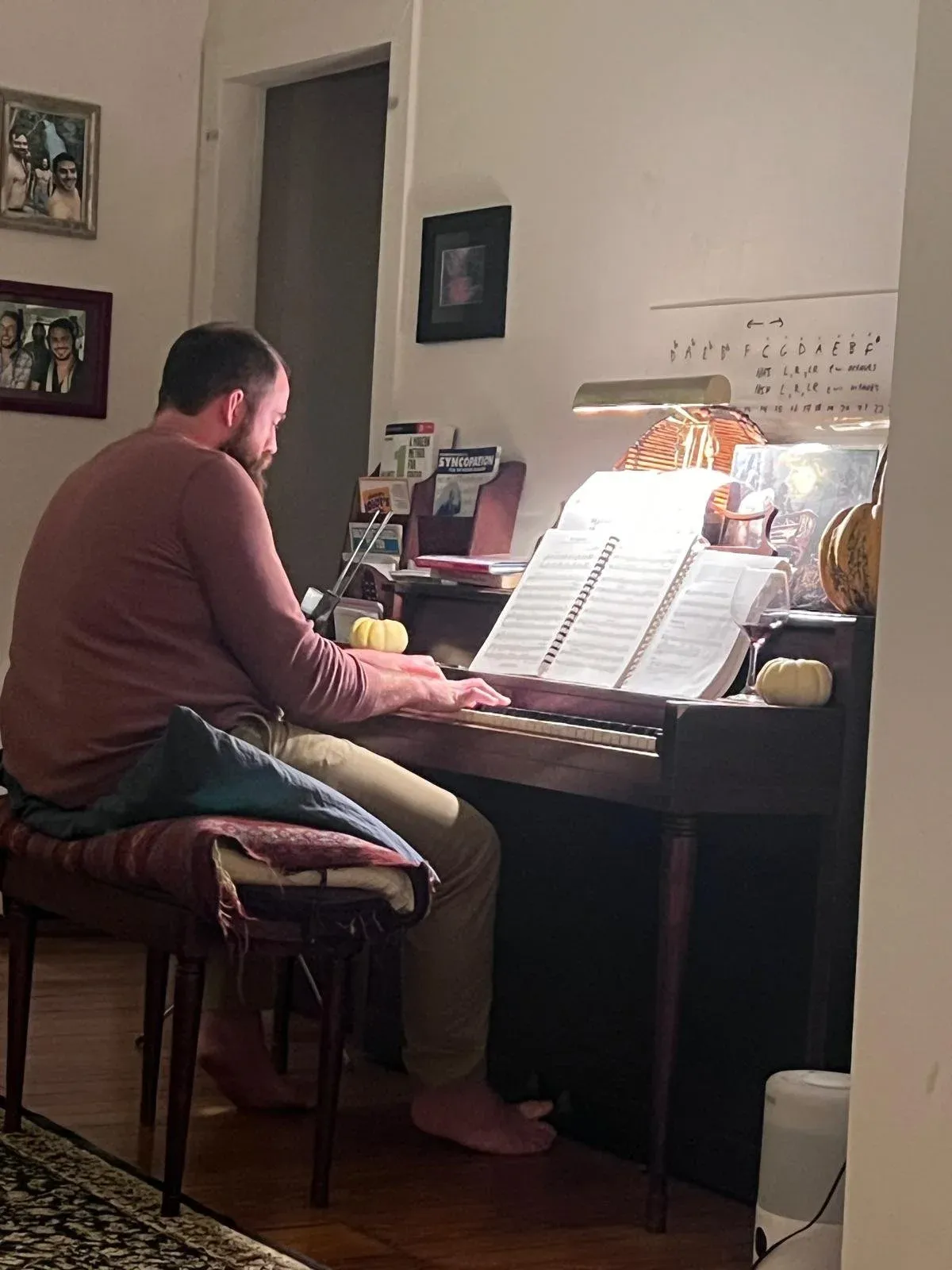 A man playing an upright piano at home, music open on the stand, framed pictures beside him.
