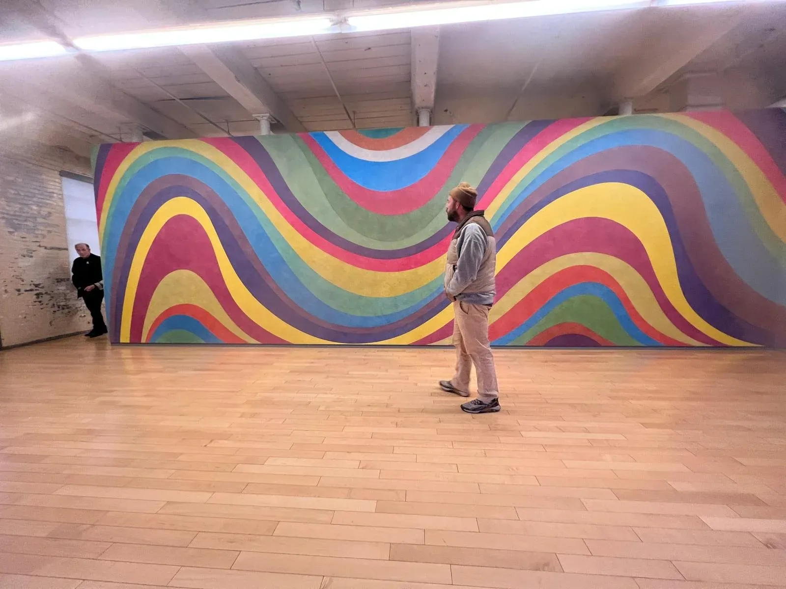 A figure standing alone in a gallery before a wall-sized mural of broad multicoloured waves.