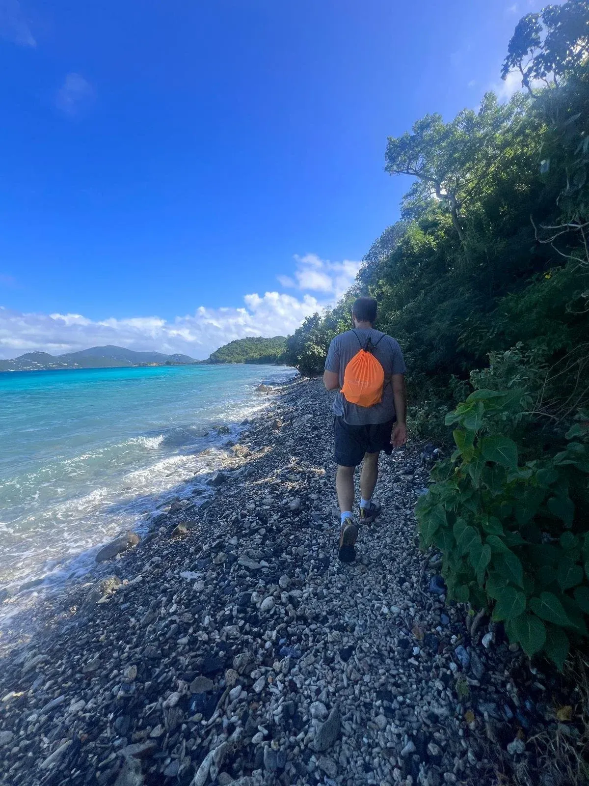 A person with an orange backpack walking a rocky shoreline above bright blue Caribbean water.