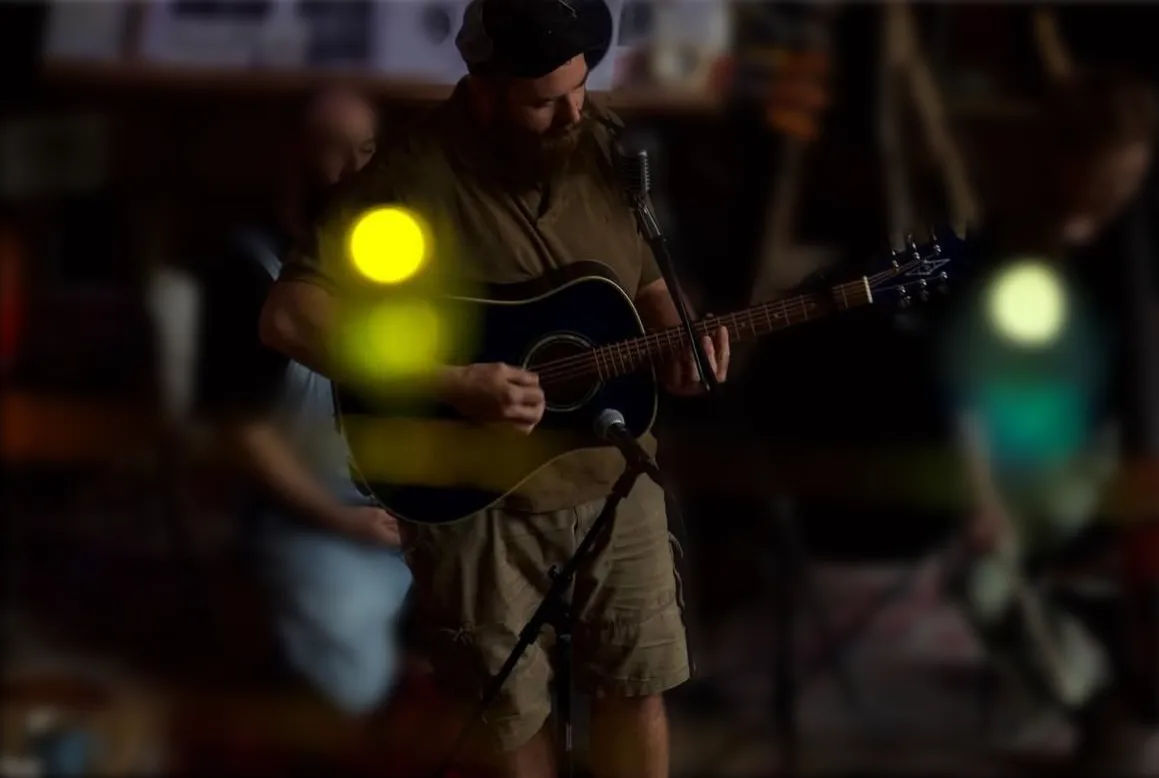 Nicholas Daly playing acoustic guitar at a microphone under colored stage lights.